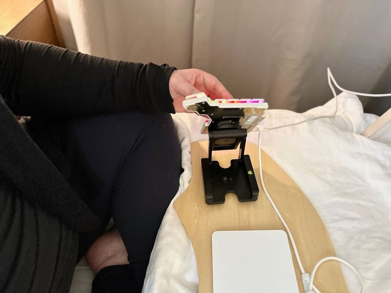 alt='Half of a split keyboard, magnetically attached to a folding stand on a wooden board, which rests on a bed covered with white linen. The stand tents the keyboard very steeply, keeping the wrist of the person using it neutral.'