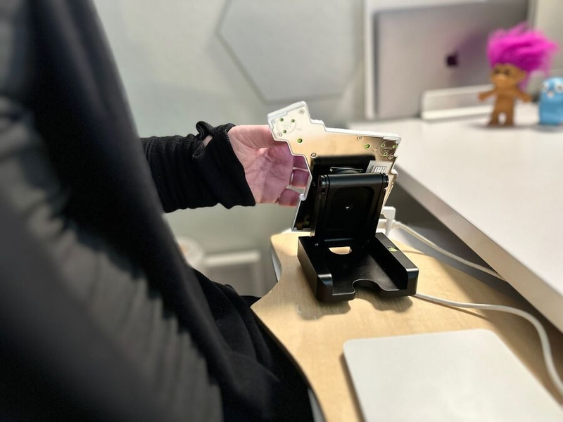 alt="Half of a split keyboard, magnetically attached to a folding stand on a wooden board, which rests on a keyboard tray on a white desk. The stand tents the keyboard very steeply, keeping the wrist of the person using it neutral."