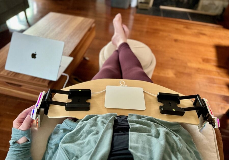 Woman on an ivory armchair, a split keyboard on a lap desk with a white trackpad between the two halves. The halves are mounted on z-shaped camera mounts, which are fully extended in the horizontal direction to enable typing without lifting the wrists from the arms of the chair.