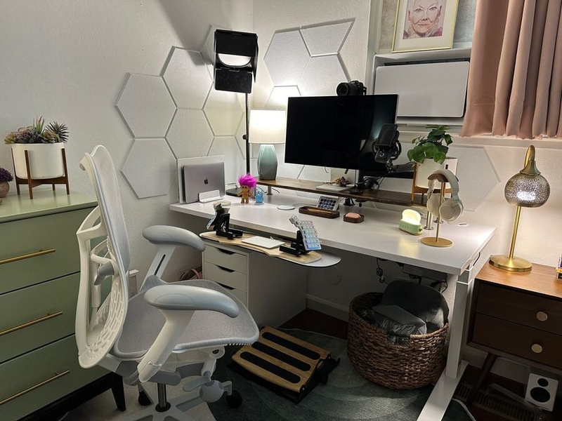 alt="White standing desk with white and grey office chair in front of it, green dresser to the left, and walnut nightstand to the right. On the desk is a monitor on a long dark wood stand along with a turquoise lamp and a leafy plant. Also on the desk are two laptops stood vertically, a few cute figurines, wooden trays for office items, and a gold headphone stand holding white headphones. The desk has a keyboard tray with a split keyboard on it, steeply tented, and a trackpad between the keyboard halves.'