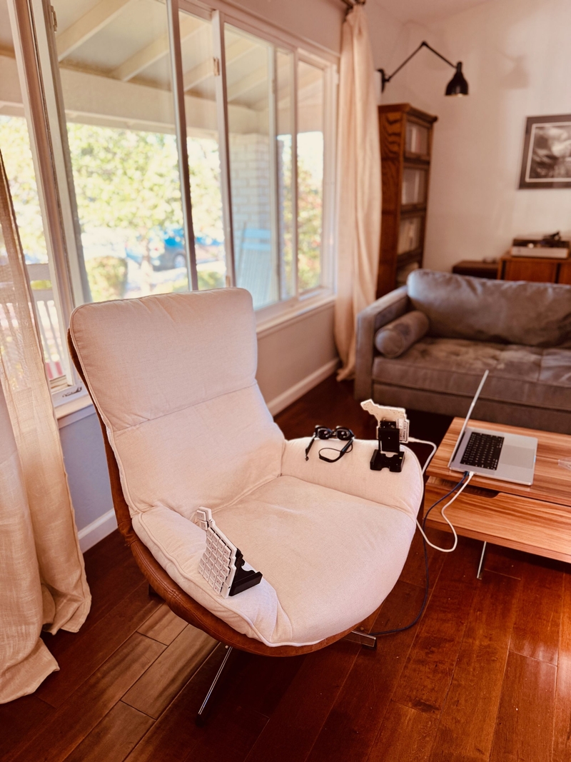 alt="Ivory-covered midcentury modern style armchair with the halves of a split keyboard resting on the arms. The chair is front of a wide window, with a coffee table and grey sofa in the background."